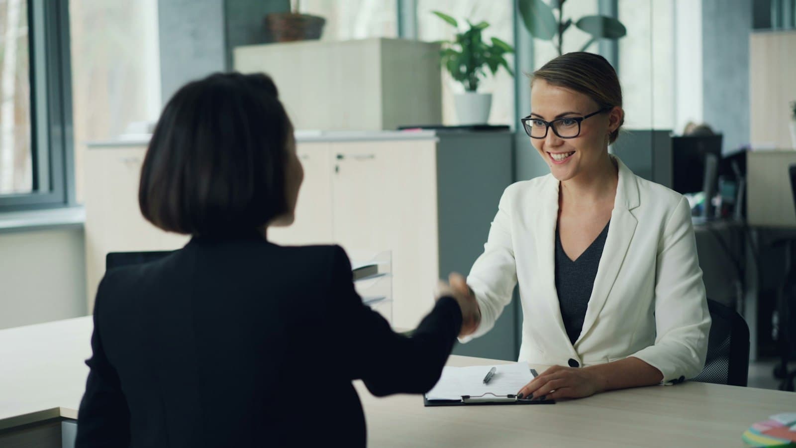 Two women shaking hands across a desk in an office