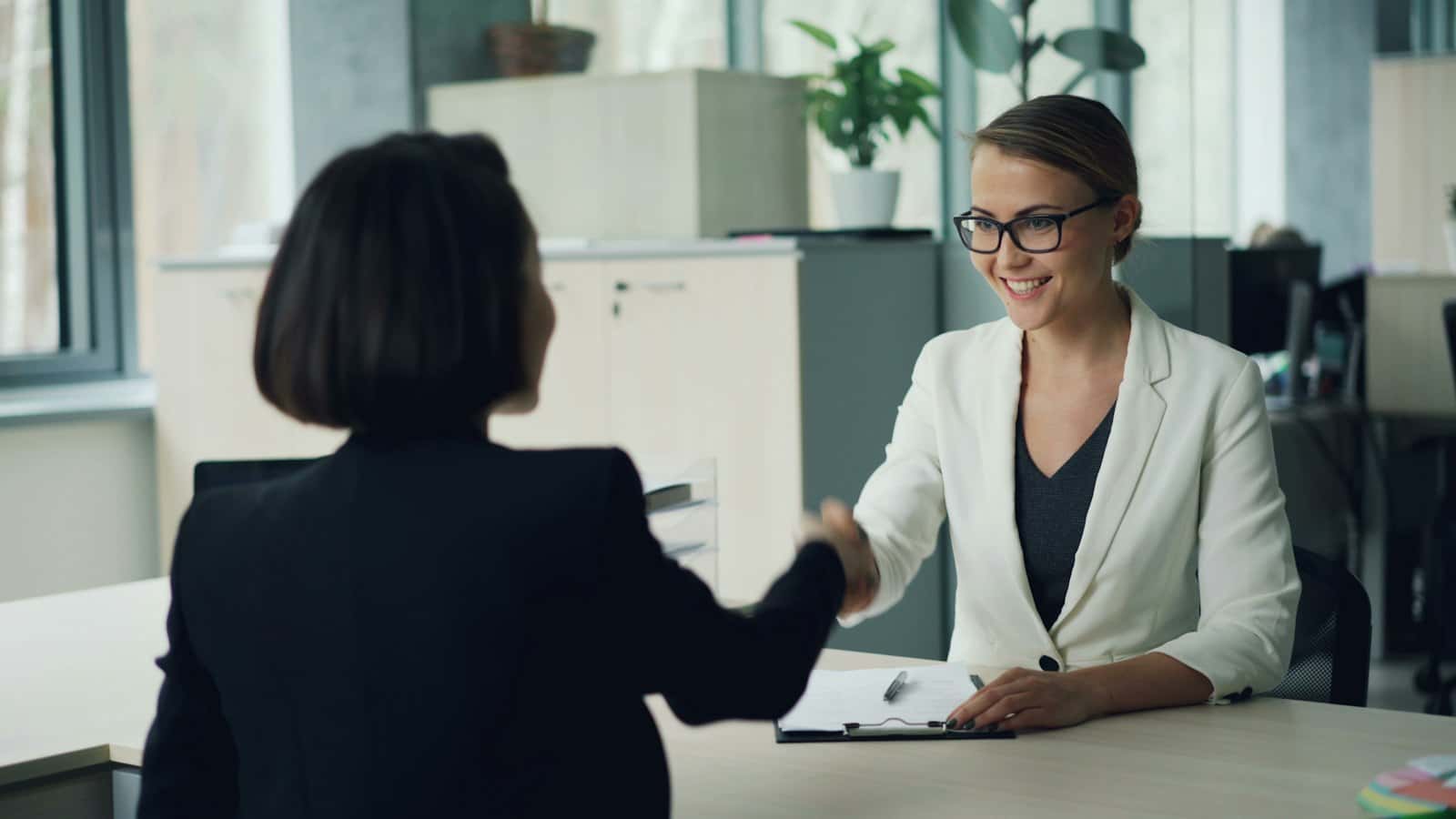 Two women shaking hands across a desk in an office